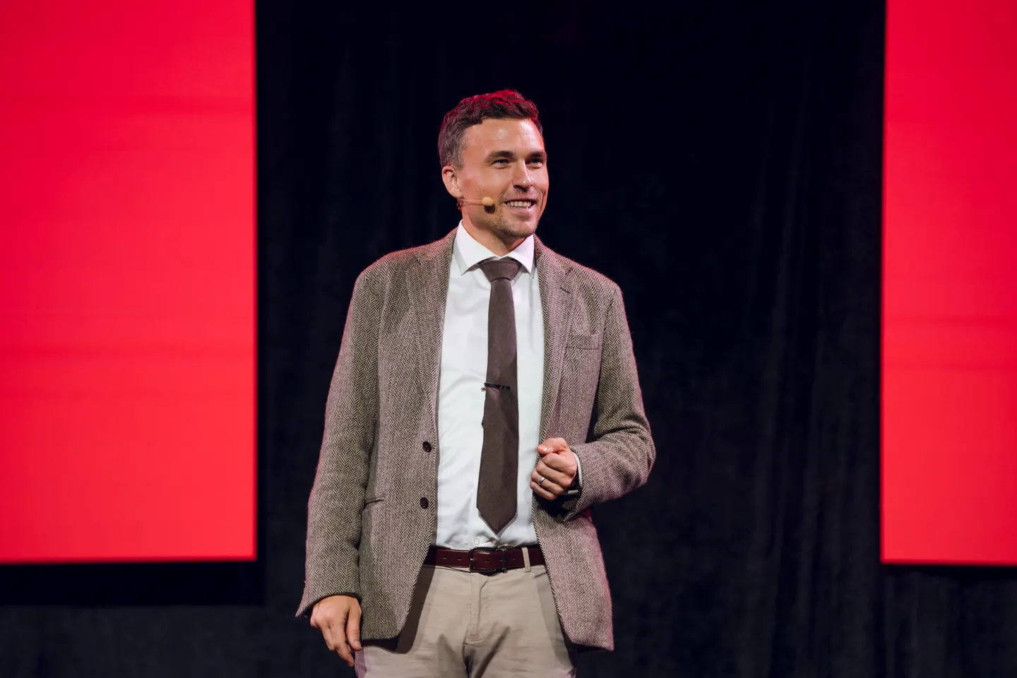 Andy Nunn speaking on stage with a headset microphone against red and black screens.